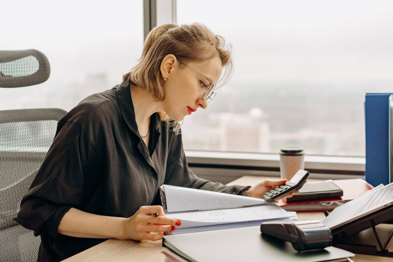 Accountant in a dark blouse working through a file of documents with a calculator at a desk beside a high-rise office window