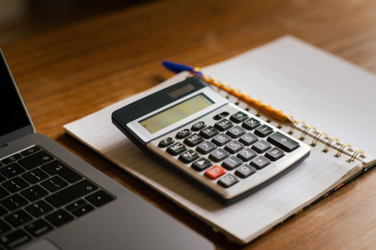 Desk calculator resting on an open spiral notebook next to a laptop on a wooden desk