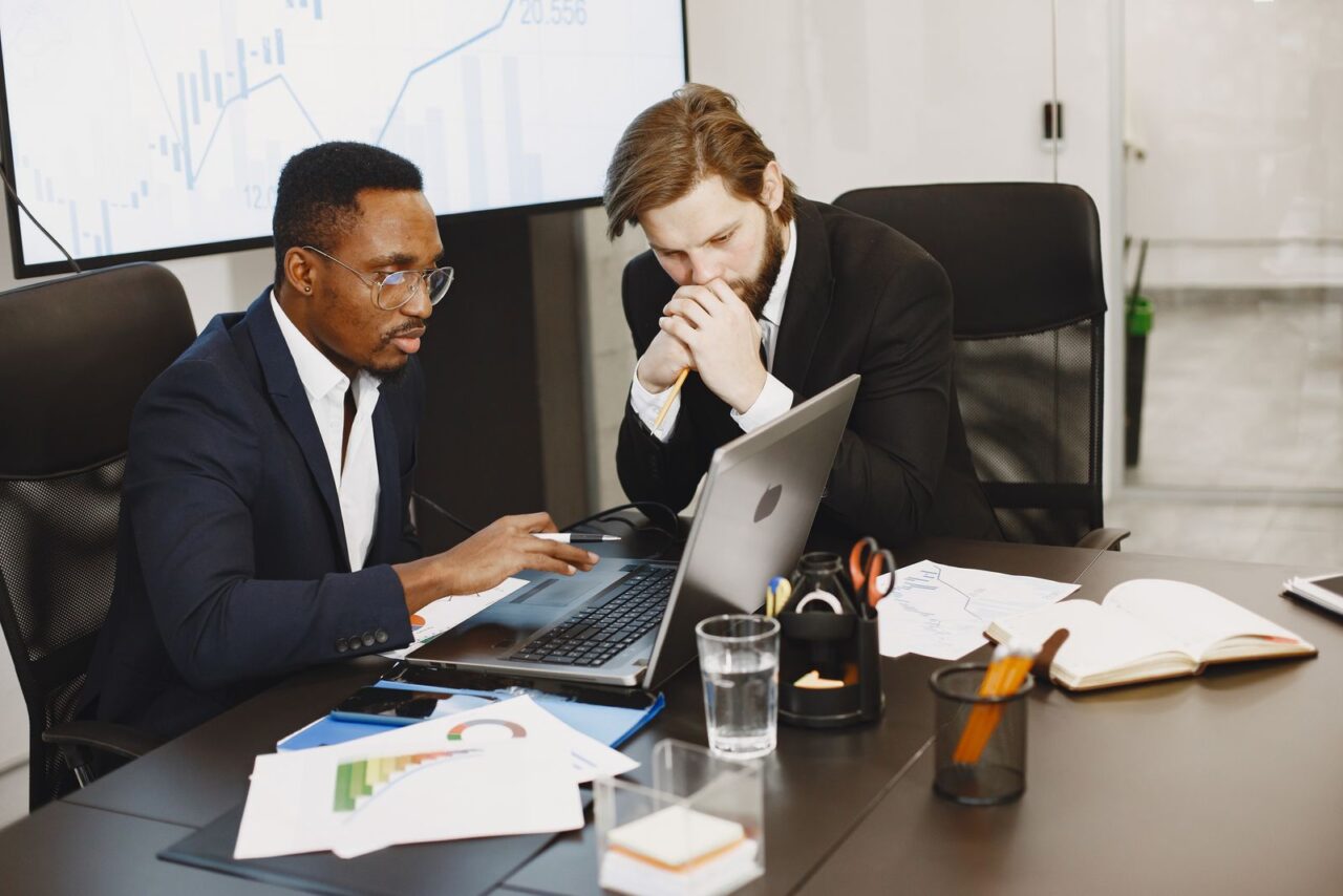 Two colleagues in suits reviewing a file on a laptop at a boardroom table