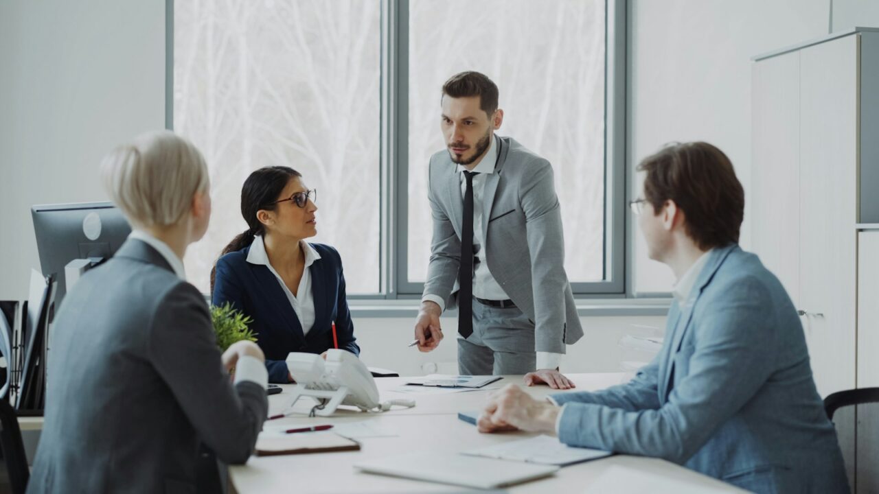 Four colleagues in business suits discussing documents around a meeting table in a bright office