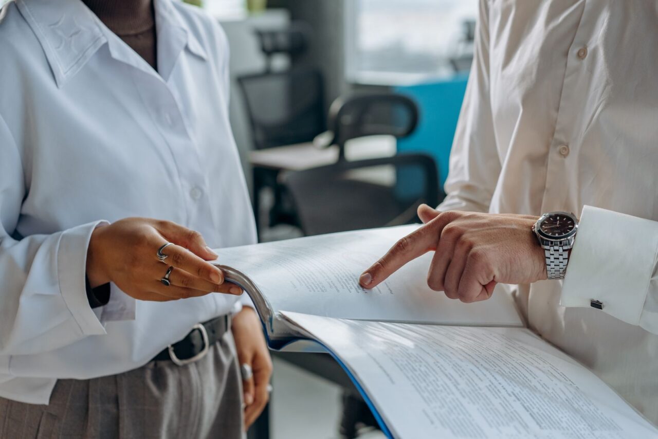 Two colleagues in white shirts reviewing a bound financial report, one pointing to a figure on the page