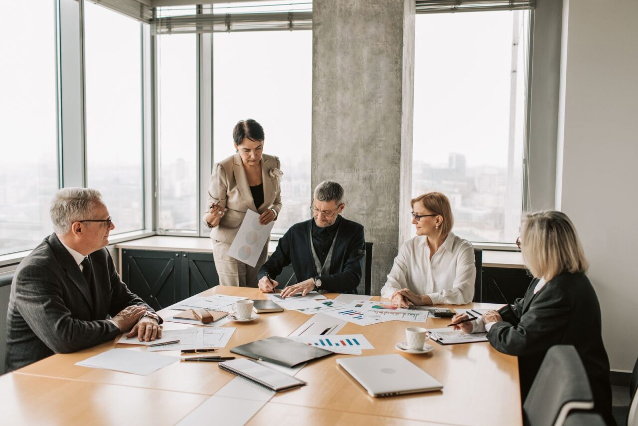 Business Valuation Services in Dubai — hero Five colleagues in business dress around a meeting table covered with printed charts and reports, beside floor-to-ceiling windows overlooking a city