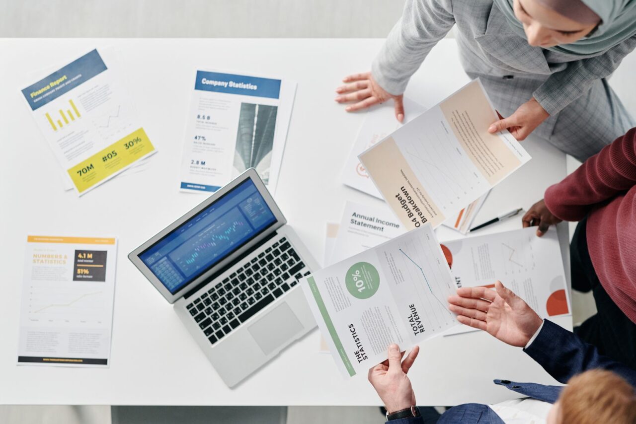Business Valuation Services in Dubai — section2 Overhead view of a white table with a laptop and printed financial reports and charts as three people review the documents