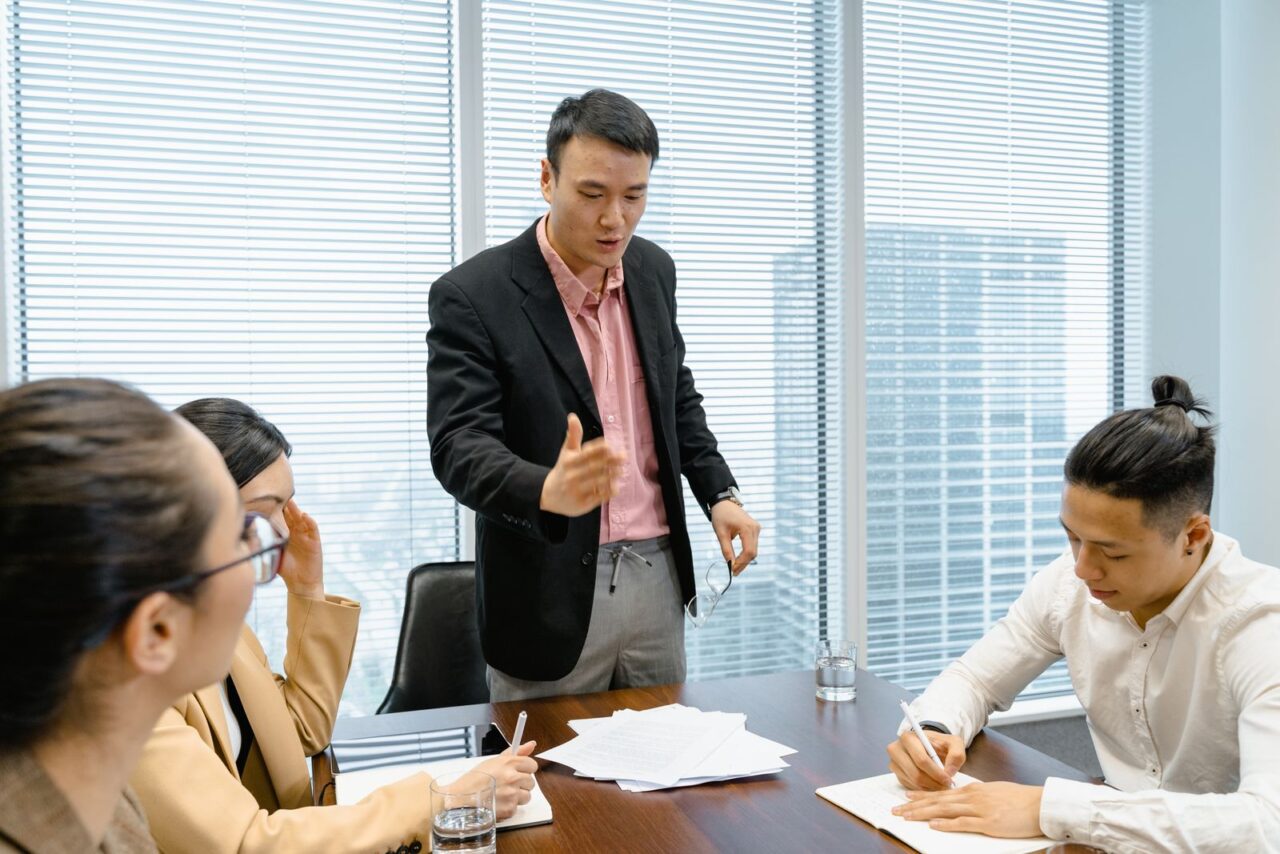 An executive standing at a boardroom table explaining a point to colleagues reviewing papers, with office towers visible through the window blinds