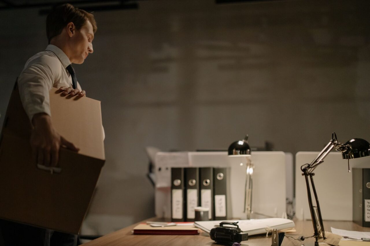 Man in a shirt and tie carrying a cardboard archive box past a desk lined with ring binders