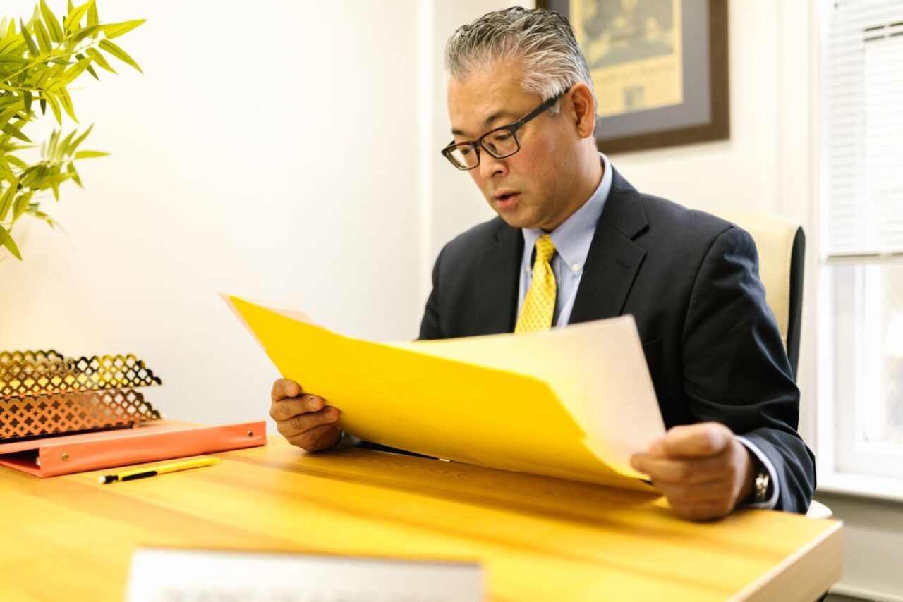 Finance professional in a suit and glasses reviewing papers from a yellow folder at his office desk