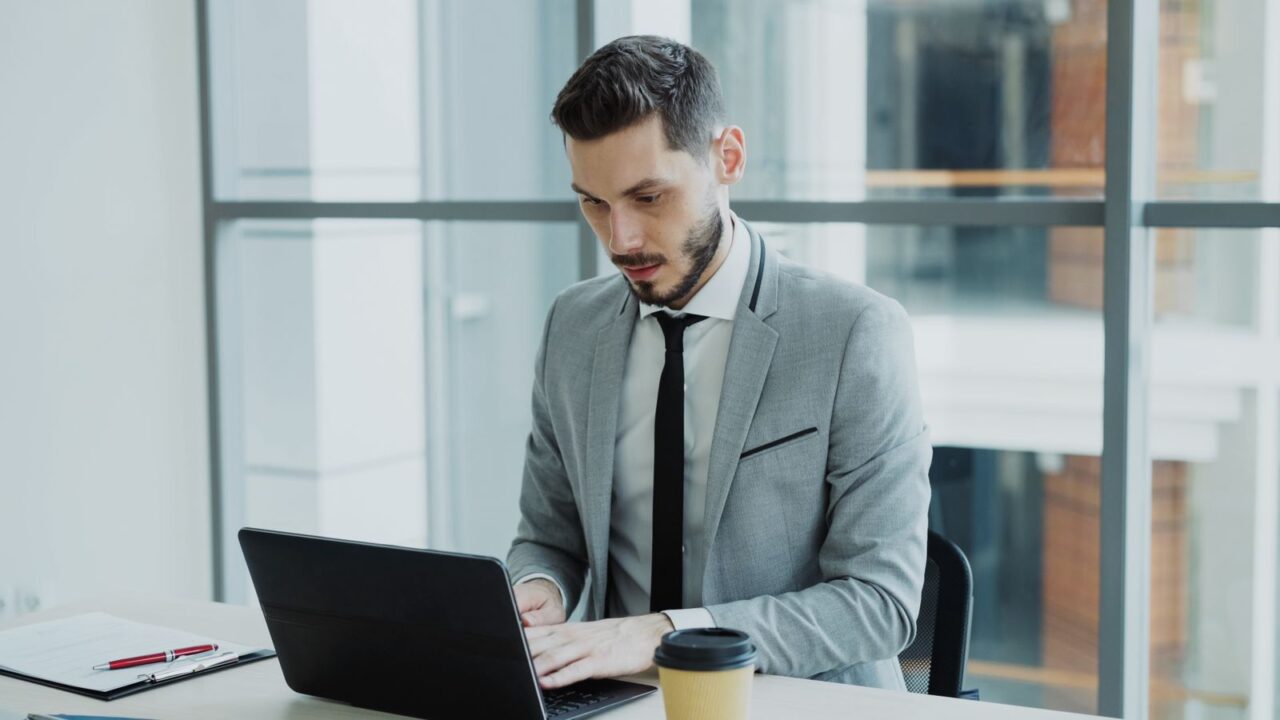 E-Invoicing in the UAE: Scope, Deadlines and Penalties — hero Man in a grey suit and tie typing on a laptop at a desk in a bright glass-walled office