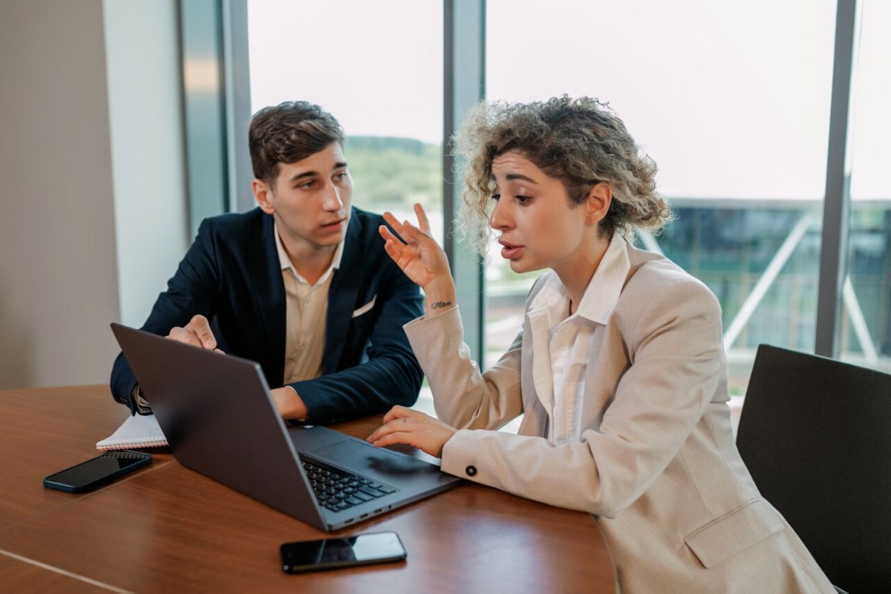 Two colleagues in business dress reviewing figures on a laptop at a meeting table beside floor-to-ceiling windows