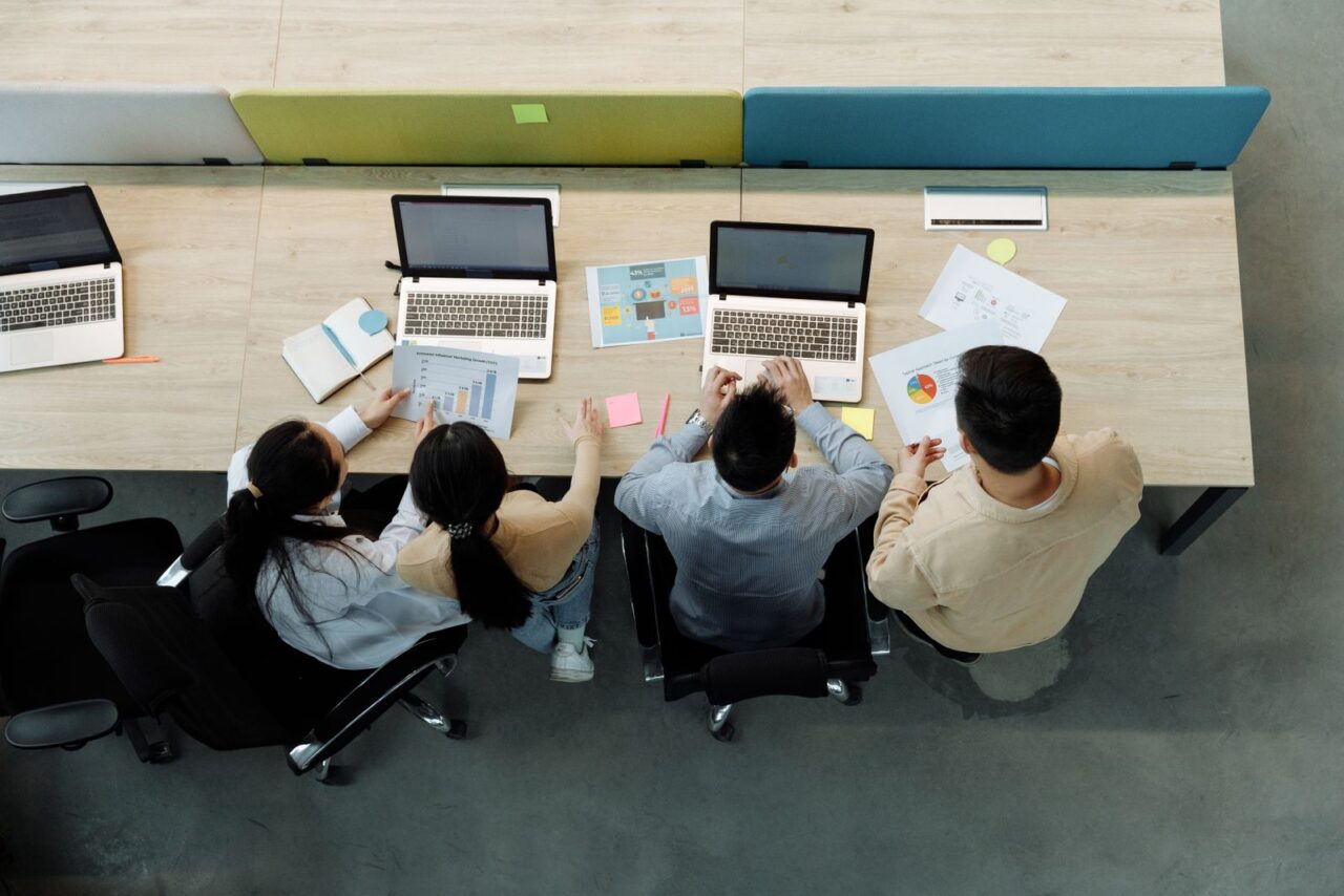 Overhead view of four colleagues working on laptops with printed charts and reports at a shared office desk