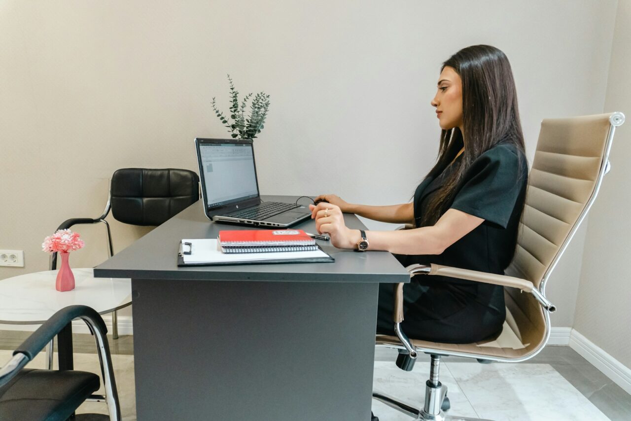 Woman in a dark blouse working on a laptop at an office desk with folders beside her
