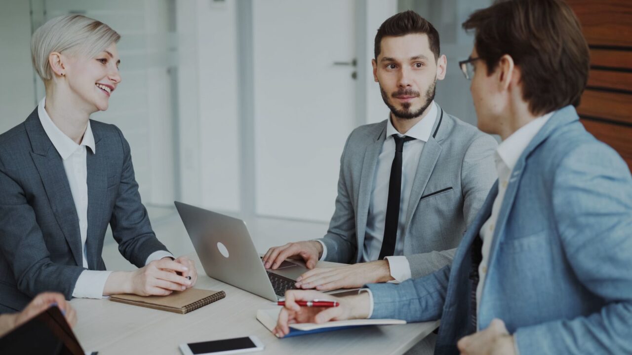 Three colleagues in business suits discussing work around a laptop at an office meeting table