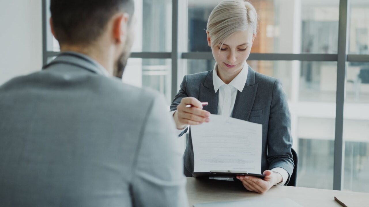 A consultant in a grey suit holds up a printed document to show a client across a desk in a bright office