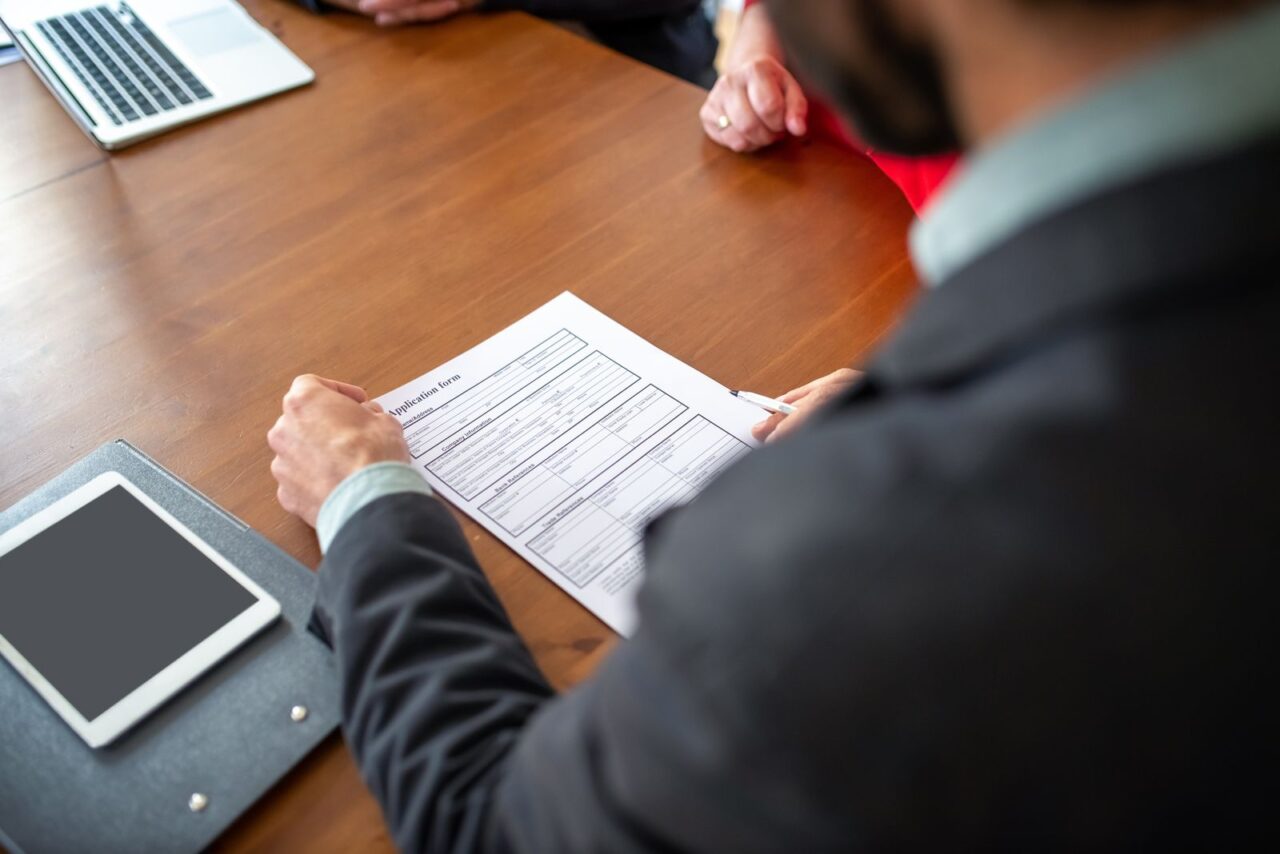 A man in a dark suit reviews a printed form at a meeting table beside a tablet and a laptop