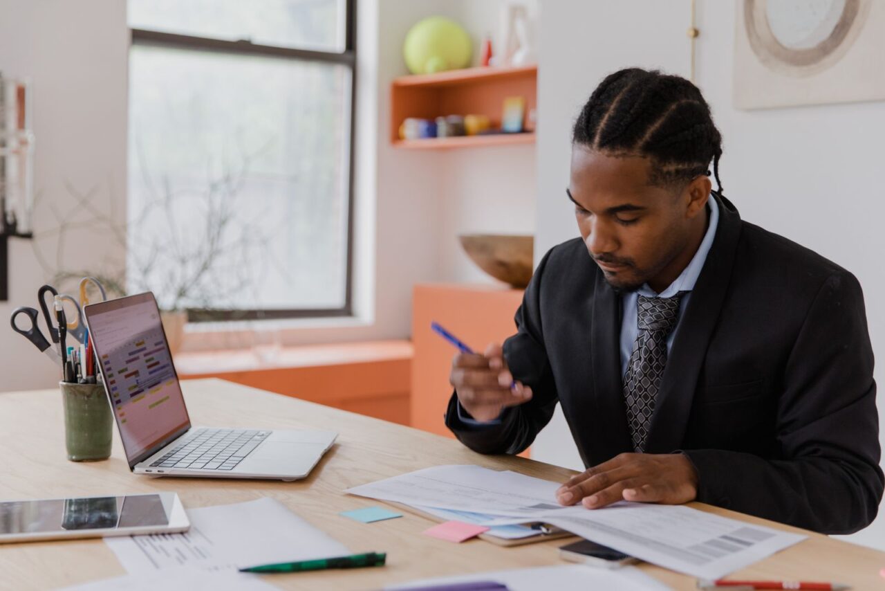 Business owner in a suit signing documents at a desk with an open laptop and tablet beside him
