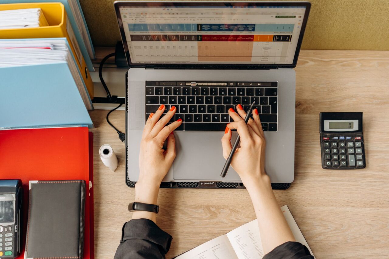 VAT Services in Dubai — hero Overhead view of hands working on a spreadsheet on a laptop, with a calculator, card terminal and folders of paperwork on the desk