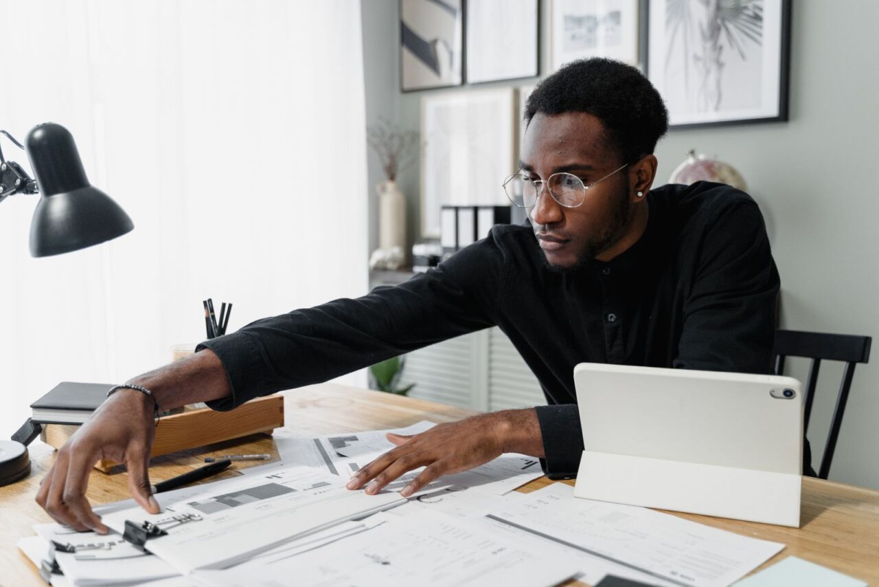 Man in glasses reviewing printed financial documents spread across his desk beside a tablet