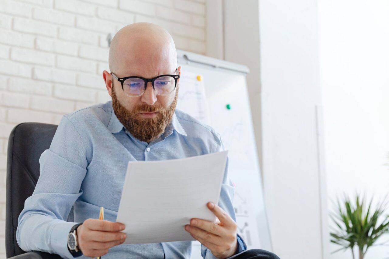 Man in glasses and a blue shirt carefully reading a printed document at his office desk